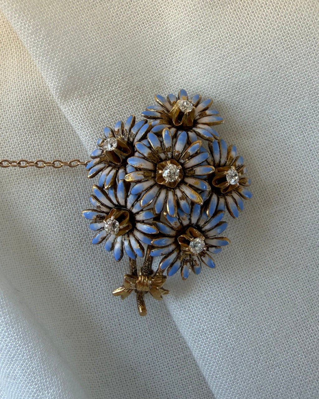 A vintage-style floral brooch or pendant with five gold-tone flower petals, each outlined in light blue enamel and set with a clear rhinestone, resting on draped off-white fabric.