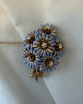 A vintage-style floral brooch or pendant with five gold-tone flower petals, each outlined in light blue enamel and set with a clear rhinestone, resting on draped off-white fabric.