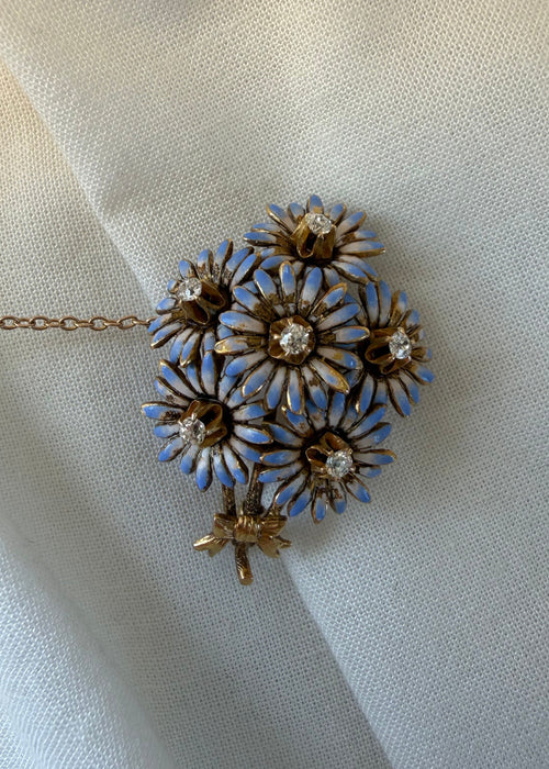 A vintage-style floral brooch or pendant with five gold-tone flower petals, each outlined in light blue enamel and set with a clear rhinestone, resting on draped off-white fabric.