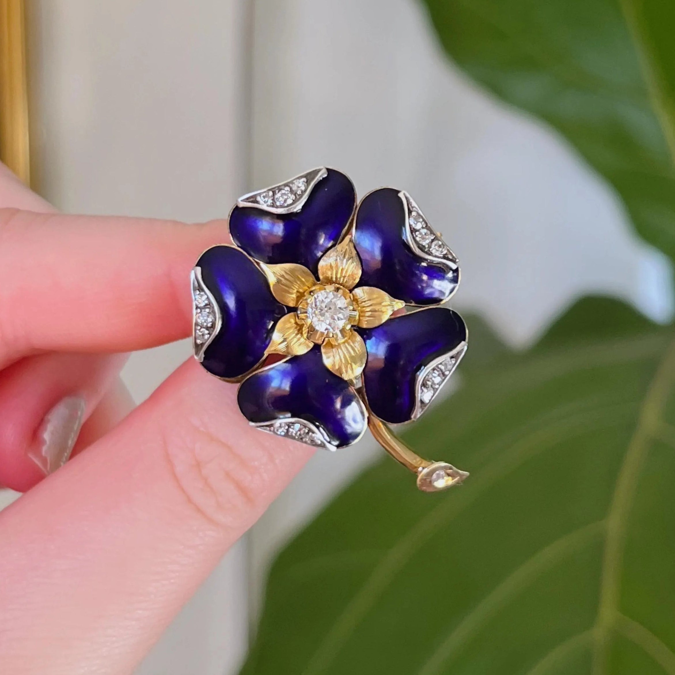 Purple flower-shaped brooch with gold center held by a hand against a green leaf background